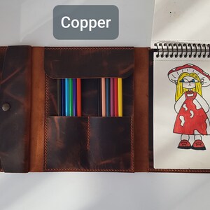 May include: A dark brown leather organizer featuring a snap closure, a central pocket holding several colored pencils, and a spiral-bound sketchbook on the right. The sketchbook page displays a drawing of a character with blonde hair and a red mushroom-patterned dress, with the text "mashrooms" and a small mushroom illustration below.