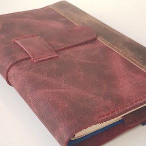 May include: A maroon leather journal cover with a distressed texture and a horizontal strap closure. The cover features visible red stitching along the edges and a darker brown leather spine section, enclosing a notebook with blue page edges.