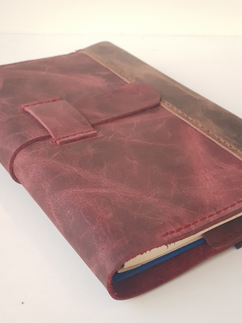 May include: A maroon leather journal cover with a distressed texture and a horizontal strap closure. The cover features visible red stitching along the edges and a darker brown leather spine section, enclosing a notebook with blue page edges.