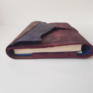 May include: A leather journal cover in deep burgundy with a dark blue flap and a single metal snap closure. The cover features visible stitching along the edges and holds a notebook with cream-colored pages, showing a blue inner lining.