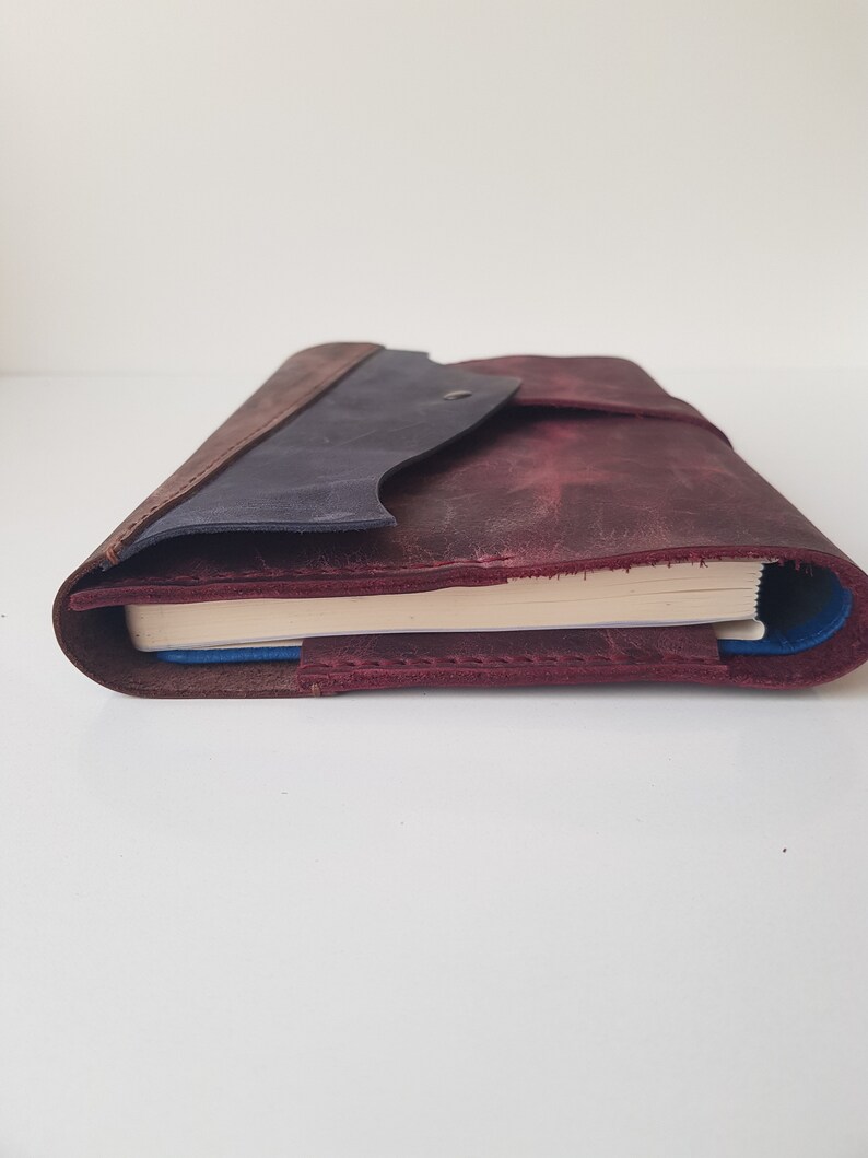 May include: A leather journal cover in deep burgundy with a dark blue flap and a single metal snap closure. The cover features visible stitching along the edges and holds a notebook with cream-colored pages, showing a blue inner lining.