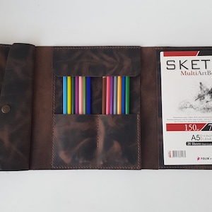 May include: A dark brown leather organizer case featuring a flap with a metal snap closure, two interior pockets holding twelve colored pencils, and an A5 MultiArtBook sketch pad with 25 sheets of 150g paper measuring 5.83 x 8.3 inches.