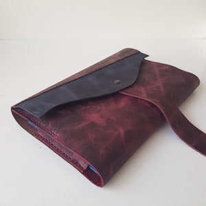 May include: A leather clutch bag featuring a deep burgundy main body with a distressed texture, a dark navy blue flap, and a single metallic snap closure. The edges are stitched with matching thread, and a thin leather strap extends from the side.