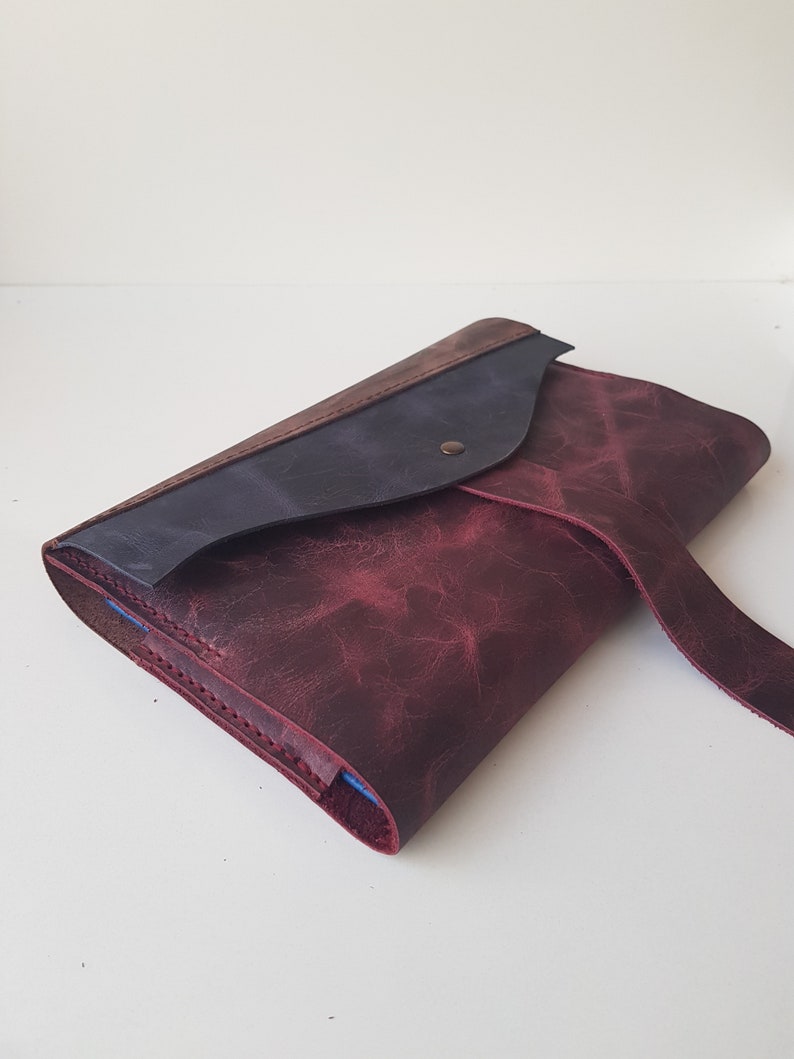 May include: A leather clutch bag featuring a deep burgundy main body with a distressed texture, a dark navy blue flap, and a single metallic snap closure. The edges are stitched with matching thread, and a thin leather strap extends from the side.