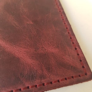 May include: A deep burgundy leather surface with a natural, mottled grain texture and visible distressed markings. The edge of the leather is secured with a straight line of matching dark red stitching, creating a clean, finished border detail.