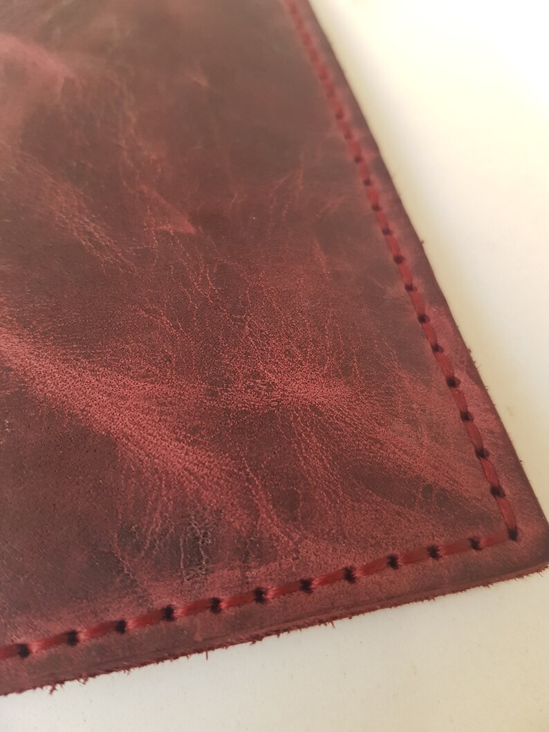 May include: A deep burgundy leather surface with a natural, mottled grain texture and visible distressed markings. The edge of the leather is secured with a straight line of matching dark red stitching, creating a clean, finished border detail.