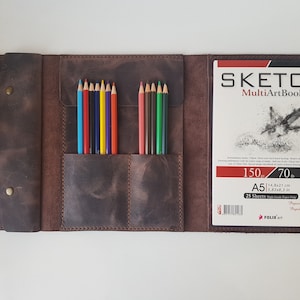 May include: A dark brown leather organizer featuring a flap with two brass snaps, two interior pockets holding ten colored pencils, and an A5 Sketch MultiArtBook with a hummingbird illustration on the cover, measuring 14.8x21 cm.
