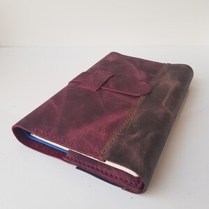 May include: A deep burgundy leather journal cover with a distressed finish and a matching leather strap closure. The cover features visible stitching along the edges and a wrap-around design that secures a notebook with cream-colored pages inside.