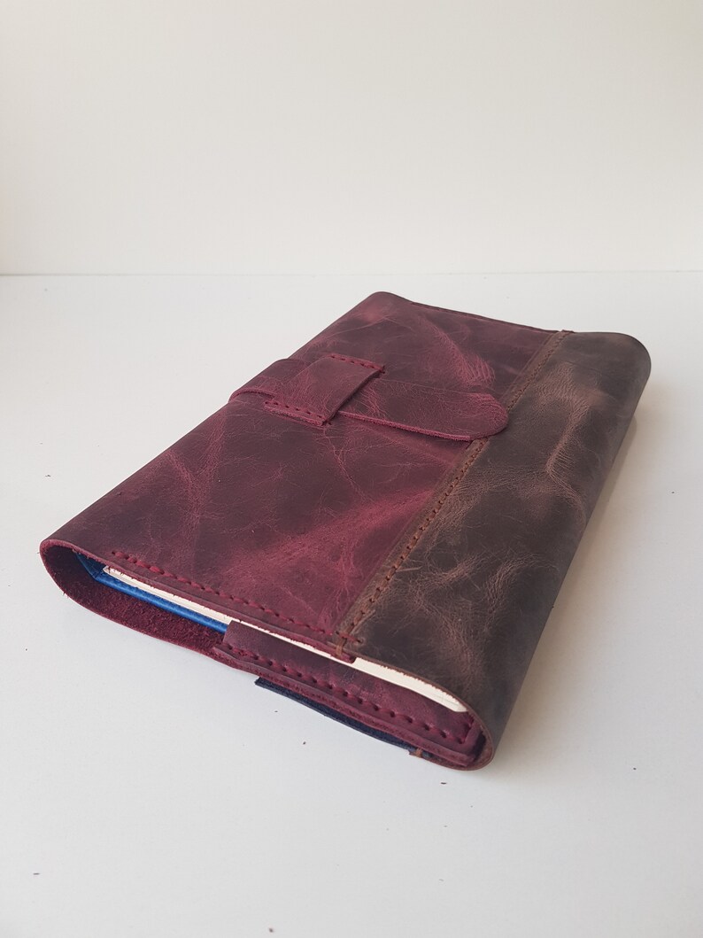 May include: A deep burgundy leather journal cover with a distressed finish and a matching leather strap closure. The cover features visible stitching along the edges and a wrap-around design that secures a notebook with cream-colored pages inside.
