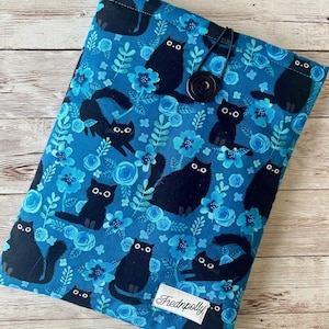 Puede incluir: Una funda de libro de tela azul con un patr&oacute;n de gato negro y flores azules. La funda tiene un cierre de bot&oacute;n y una etiqueta negra con el texto "Frednpolly".