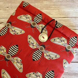 May include: Red fabric pouch with a black drawstring closure and a gold button. The pouch is decorated with a repeating pattern of embroidered bees with black and gold details.