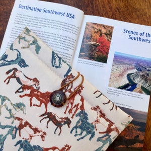 Puede incluir: Primer plano de una cubierta de libro de tela con un patrón de caballos en tonos marrones, azules y verdes. La cubierta está doblada sobre un libro titulado "Destination Southwest USA" e incluye imágenes de cañones y ríos.