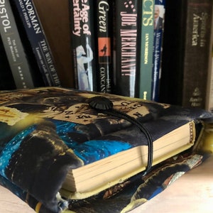 Op de afbeelding: Een zwarte boekomslag met een kleurrijk ontwerp met een blauw, geel en wit patroon. De omslag heeft een zwarte knop en een zwarte band om het boek gesloten te houden.