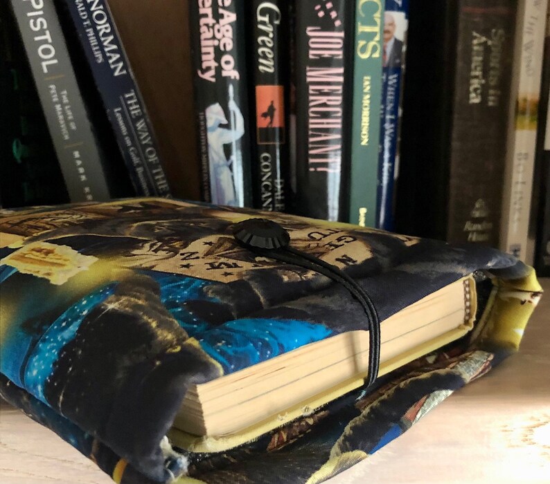 Op de afbeelding: Een zwarte boekomslag met een kleurrijk ontwerp met een blauw, geel en wit patroon. De omslag heeft een zwarte knop en een zwarte band om het boek gesloten te houden.
