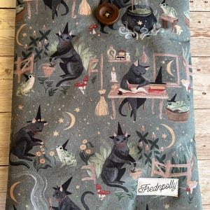 May include: A book sleeve featuring a repeating pattern of black cats in witch hats, frogs, moons, stars, and cauldrons on a grey-green fabric. It has a button closure and a label reading "Frednpolly".