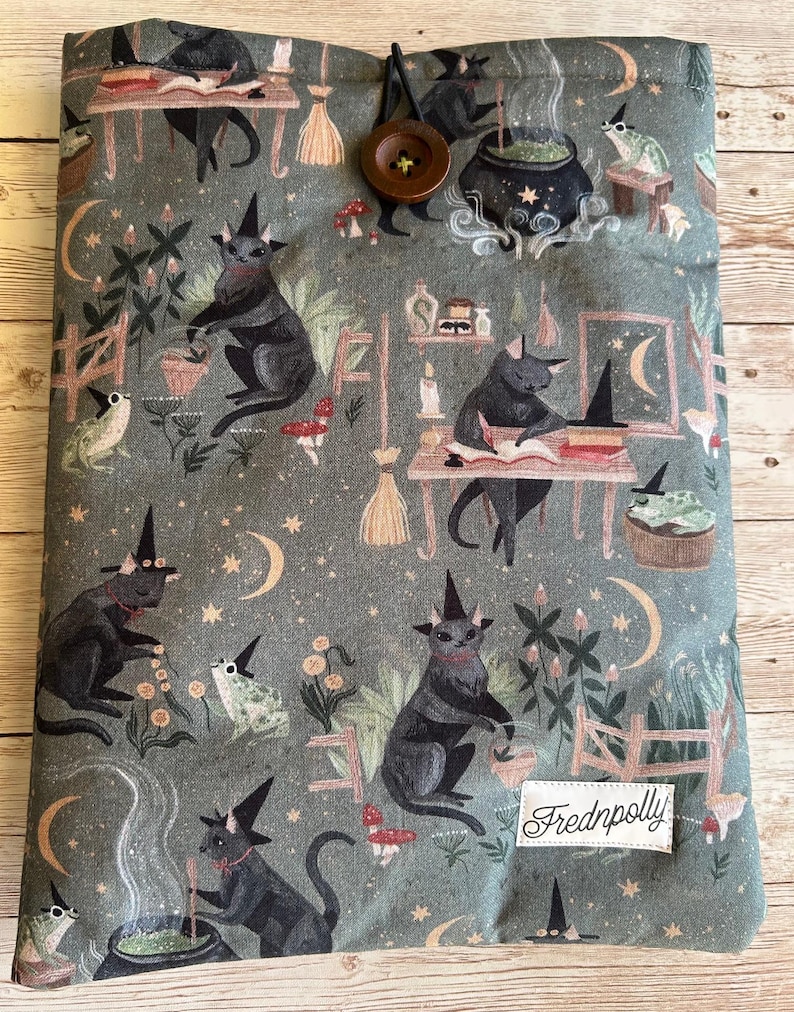 May include: A book sleeve featuring a repeating pattern of black cats in witch hats, frogs, moons, stars, and cauldrons on a grey-green fabric. It has a button closure and a label reading "Frednpolly".