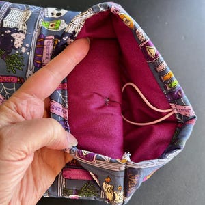 May include: A fabric pouch with a dark purple interior and a gray exterior patterned with witch-themed illustrations. The exterior design includes images of books, candles, and potion bottles. A white elastic cord is visible inside the pouch.