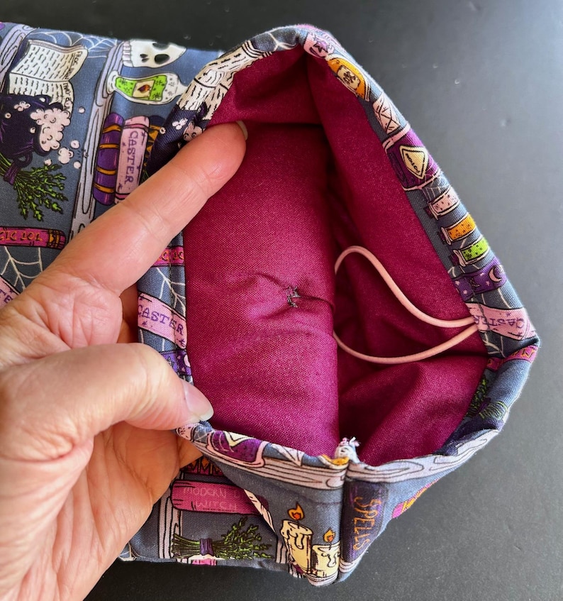 May include: A fabric pouch with a dark purple interior and a gray exterior patterned with witch-themed illustrations. The exterior design includes images of books, candles, and potion bottles. A white elastic cord is visible inside the pouch.