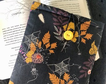 Könnte beinhalten: Eine schwarze Buchhülle mit einem Blumen- und Spinnennetz-Muster. Das Design umfasst orange, gelbe und lila Blumen und Blätter. Ein gelber Knopf und eine schwarze Kordel sichern die Hülle. Ein offenes Buch ist dahinter zu sehen.