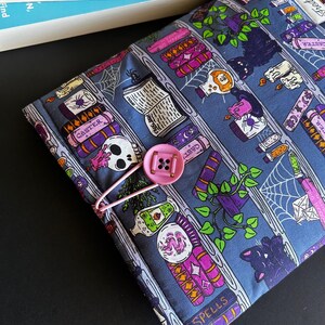 May include: A blue fabric book cover with a pink button closure. The cover features a repeating pattern of spell books, potion bottles, skulls, and spiderwebs. The cover is designed to hold a book.