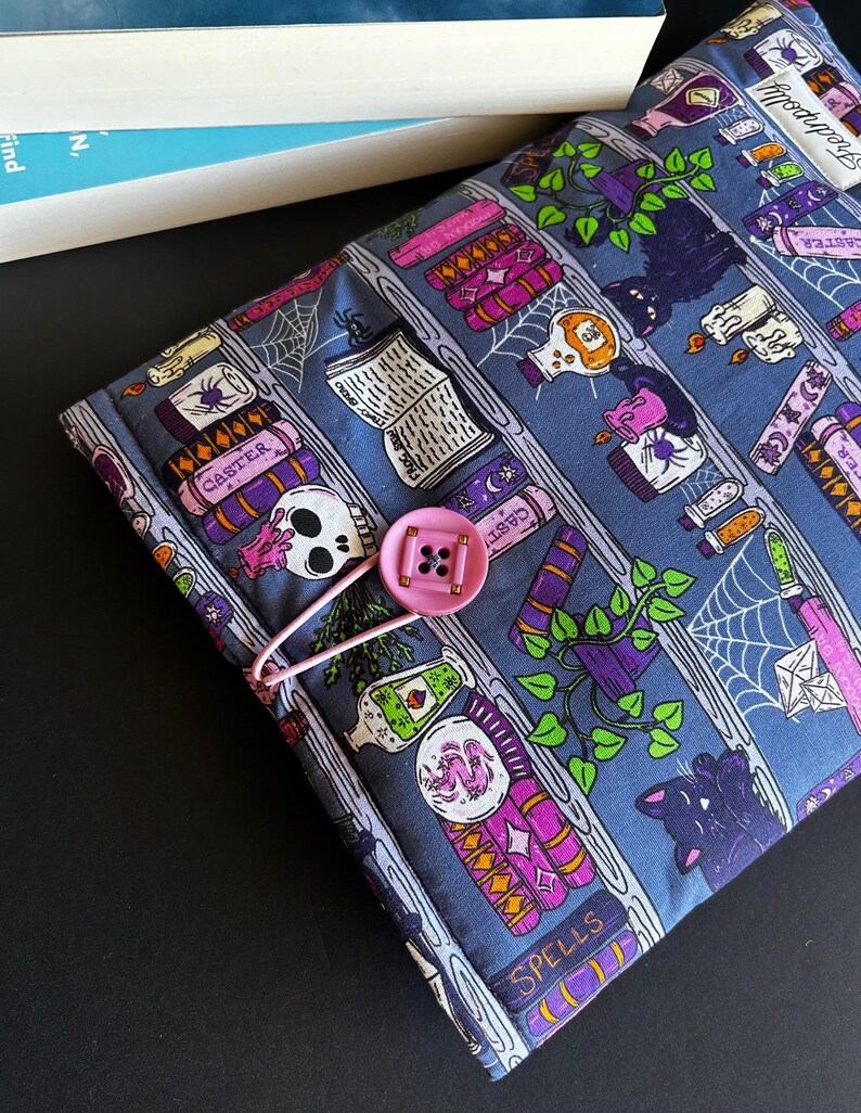 May include: A blue fabric book cover with a pink button closure. The cover features a repeating pattern of spell books, potion bottles, skulls, and spiderwebs. The cover is designed to hold a book.