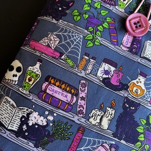 May include: A blue fabric item with a repeating pattern of shelves filled with witchy items. The shelves hold potion bottles, spell books, candles, and black cats. A pink button and loop closure is visible on the right side.