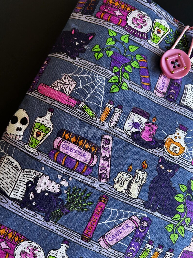 May include: A blue fabric item with a repeating pattern of shelves filled with witchy items. The shelves hold potion bottles, spell books, candles, and black cats. A pink button and loop closure is visible on the right side.