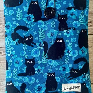 Puede incluir: Una funda de libro de tela azul con cierre de bot&oacute;n negro. La funda presenta un patr&oacute;n repetido de gatos negros y flores azules. El texto "Frednpolly" est&aacute; impreso en la esquina inferior derecha de la funda.