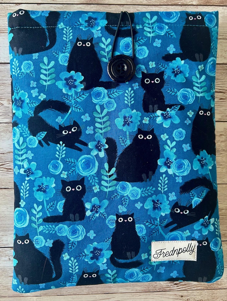 Puede incluir: Una funda de libro de tela azul con cierre de bot&oacute;n negro. La funda presenta un patr&oacute;n repetido de gatos negros y flores azules. El texto "Frednpolly" est&aacute; impreso en la esquina inferior derecha de la funda.