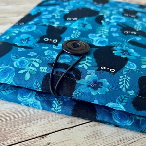 Puede incluir: Una bolsa de tela azul con cierre de bot&oacute;n negro y estampado de gato negro. La tela tiene un patr&oacute;n floral con flores y hojas azules.