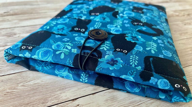 Puede incluir: Una bolsa de tela azul con cierre de bot&oacute;n negro y estampado de gato negro. La tela tiene un patr&oacute;n floral con flores y hojas azules.