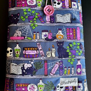 May include: A blue fabric book cover with a whimsical bookshelf design. The cover features shelves with books, potion bottles, candles, and black cats. A pink button and loop closure is at the top. The brand name "Frednpolly" is on a white label.