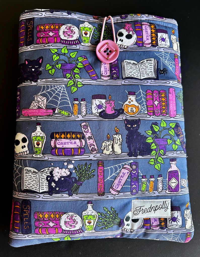 May include: A blue fabric book cover with a whimsical bookshelf design. The cover features shelves with books, potion bottles, candles, and black cats. A pink button and loop closure is at the top. The brand name "Frednpolly" is on a white label.