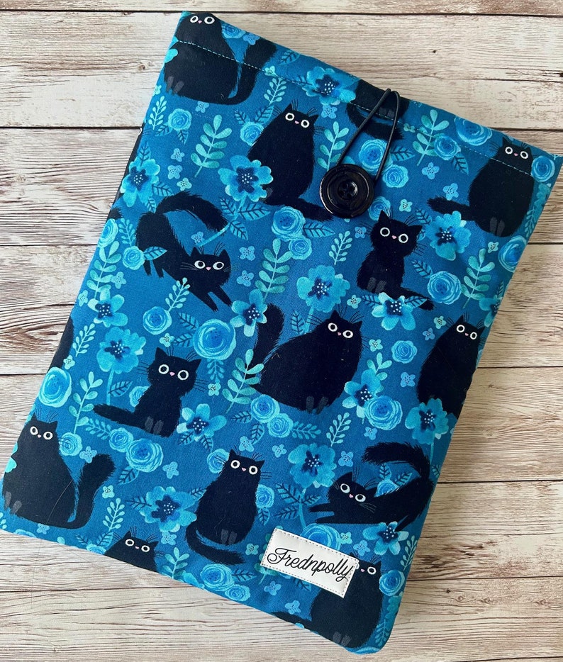 Puede incluir: Una funda de libro de tela azul con un patr&oacute;n de gato negro y flores azules. La funda tiene un cierre de bot&oacute;n y una etiqueta negra con el texto "Frednpolly".