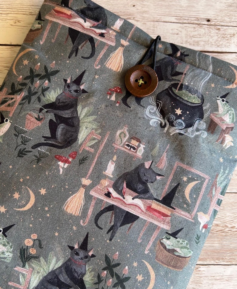 May include: A grey fabric book sleeve with a pattern of black cats in witch hats, cauldrons, and crescent moons. It has a brown button closure and a loop. The design has a witchy, magical theme, with a button closure.
