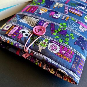 May include: A blue fabric book sleeve with a pink button closure. The sleeve features a repeating pattern of books, potion bottles, skulls, and spiderwebs. The design includes purple, pink, and white accents. The book sleeve is designed to protect books.