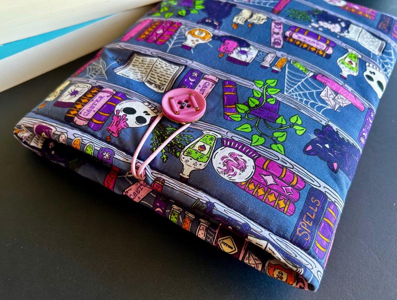 May include: A blue fabric book sleeve with a pink button closure. The sleeve features a repeating pattern of books, potion bottles, skulls, and spiderwebs. The design includes purple, pink, and white accents. The book sleeve is designed to protect books.