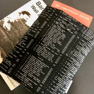 Pode incluir: Capa de livro em preto e branco com fecho de botão. O tecido apresenta uma impressão vintage de texto e ilustrações científicas, com as palavras "60th Anniversary Edition" visíveis.