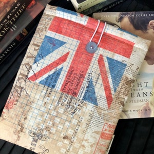 Puede incluir: Una funda para libros con un diseño de la Union Jack. La funda está hecha de tela con un patrón de cuadrícula y tiene un cierre de botón. El texto "Joseph Foster & Sons" es visible en la funda.