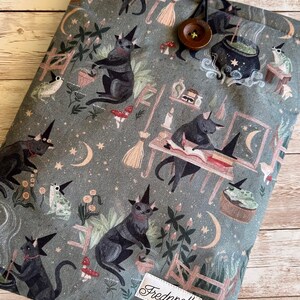 May include: A grey fabric book sleeve with a repeating pattern of black cats in witch hats, moons, stars, and mushrooms. It has a button closure and a label reading "Frednpolly".