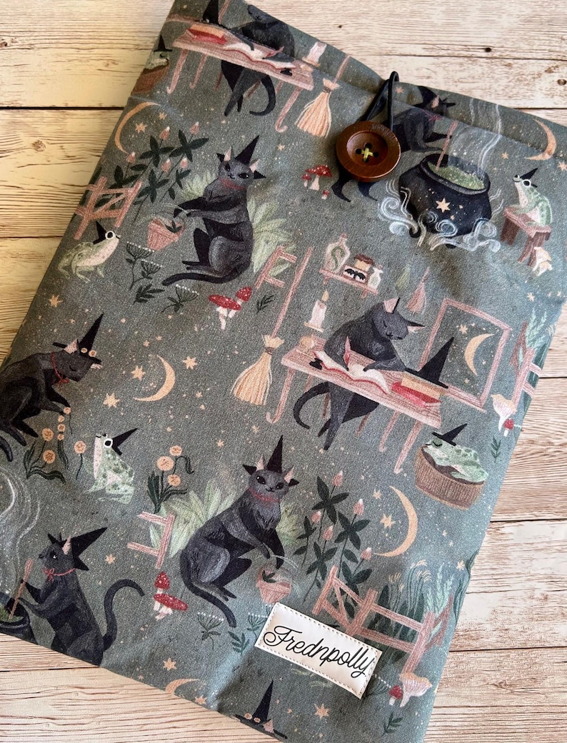 May include: A grey fabric book sleeve with a repeating pattern of black cats in witch hats, moons, stars, and mushrooms. It has a button closure and a label reading "Frednpolly".