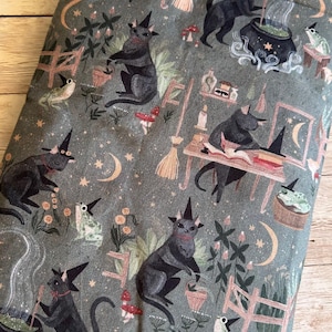 May include: A fabric item featuring a repeating pattern of black cats in witch hats, frogs, mushrooms, and crescent moons on a grey-green backdrop. The cats are shown in different activities, including stirring cauldrons and reading books. The design has a Halloween theme.