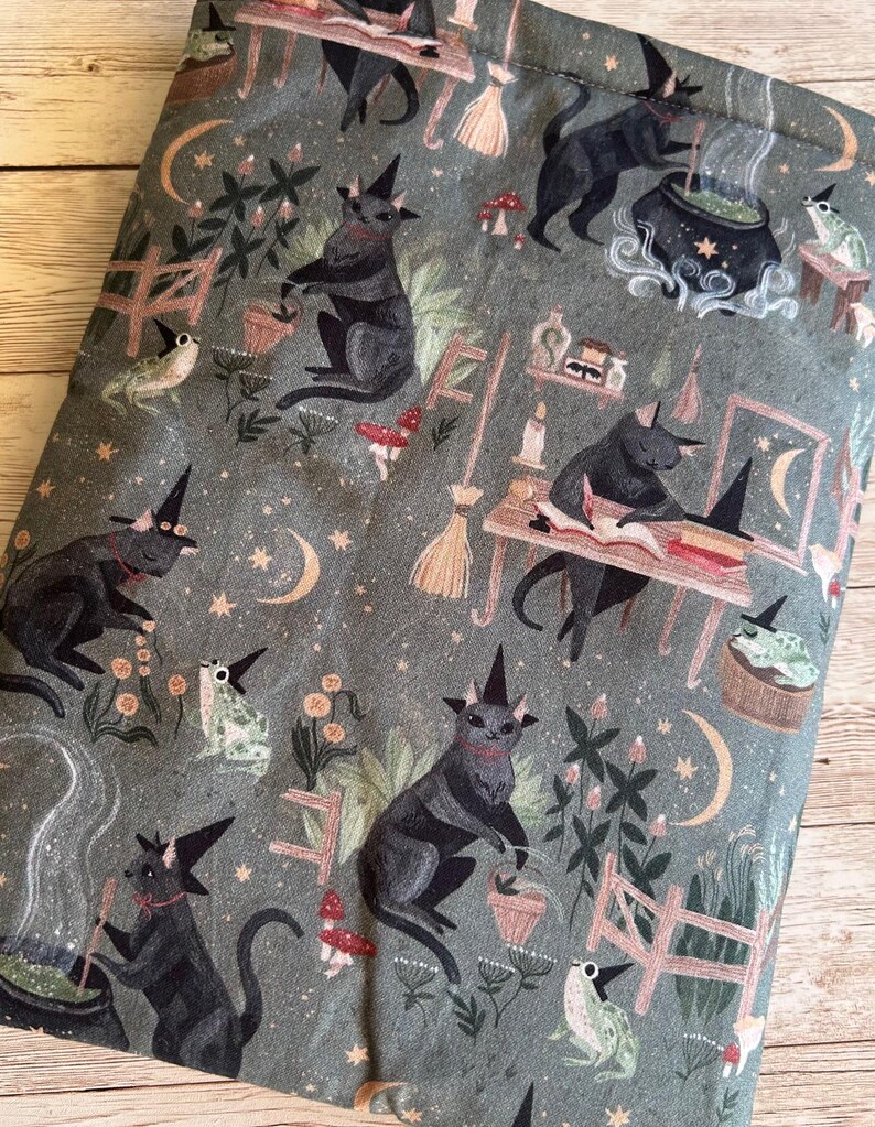 May include: A fabric item featuring a repeating pattern of black cats in witch hats, frogs, mushrooms, and crescent moons on a grey-green backdrop. The cats are shown in different activities, including stirring cauldrons and reading books. The design has a Halloween theme.