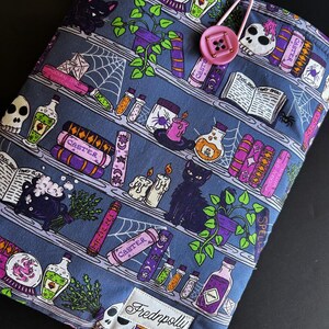 May include: A blue fabric book sleeve featuring a whimsical bookshelf design with potion bottles, spell books, and a black cat. The sleeve has a pink button closure and the brand name "Frednpolly" on a white label.