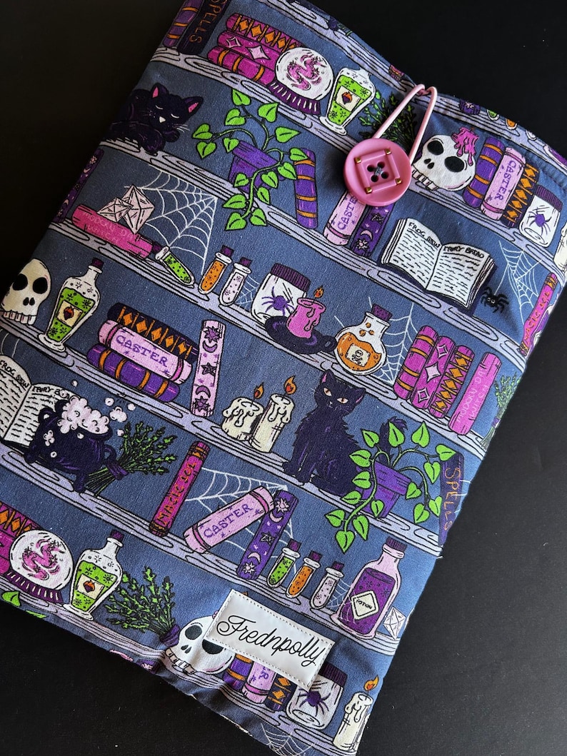 May include: A blue fabric book sleeve featuring a whimsical bookshelf design with potion bottles, spell books, and a black cat. The sleeve has a pink button closure and the brand name "Frednpolly" on a white label.