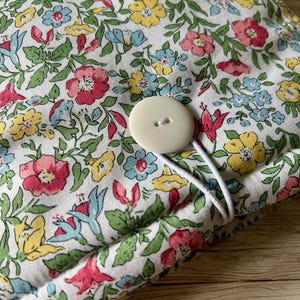 Op de afbeelding: Een witte stoffen tas met een bloemenpatroon in tinten roze, geel, blauw en groen. De tas heeft een witte knoop en een witte elastische band.