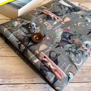 May include: A grey fabric book sleeve with a pattern of witches, bats, brooms, and moons. It has a wooden button and elastic loop closure. The sleeve is designed to protect books. The measurements are in centimetres.