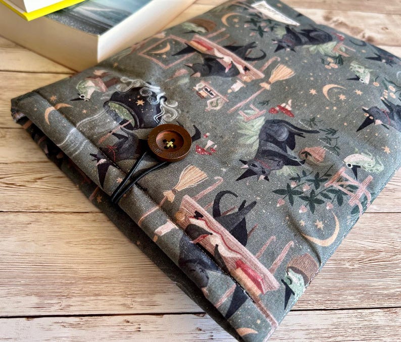 May include: A grey fabric book sleeve with a pattern of witches, bats, brooms, and moons. It has a wooden button and elastic loop closure. The sleeve is designed to protect books. The measurements are in centimetres.