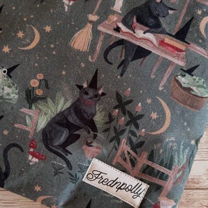 May include: Dark green fabric with a repeating pattern of black cats in witch hats, moons, stars, and mushrooms. A small wooden fence and the label "Frednpolly" are also visible.