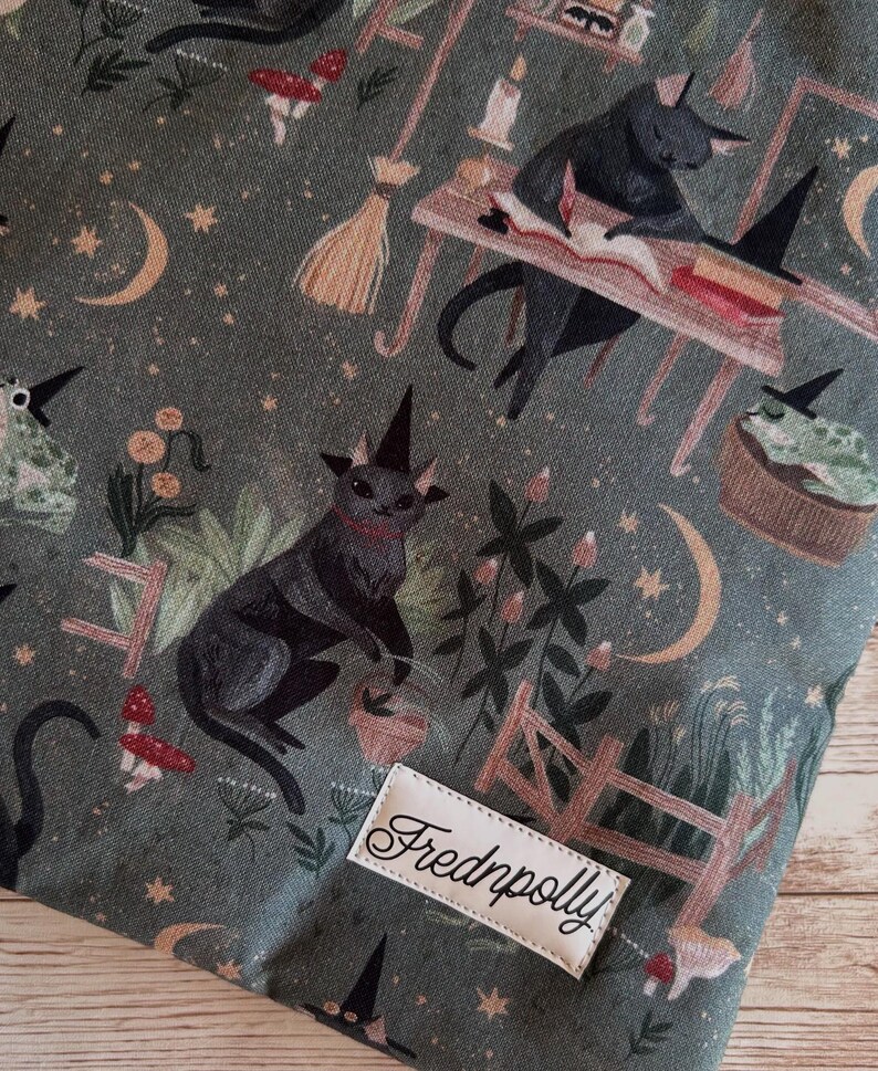 May include: Dark green fabric with a repeating pattern of black cats in witch hats, moons, stars, and mushrooms. A small wooden fence and the label "Frednpolly" are also visible.